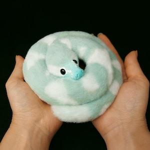 May include: A plush snake toy featuring a soft, fuzzy texture with a pattern of alternating mint green and white patches. The snake has a smooth, light blue molded plastic head with dark, round eyes and a coiled body shape.