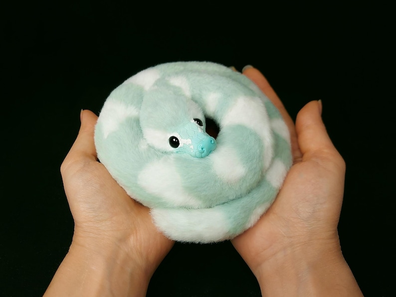 May include: A plush snake toy featuring a soft, fuzzy texture with a pattern of alternating mint green and white patches. The snake has a smooth, light blue molded plastic head with dark, round eyes and a coiled body shape.