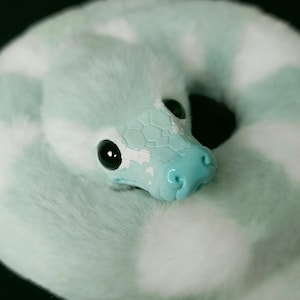 May include: A plush snake toy coiled in a circle, featuring soft mint green and white faux fur. The head is crafted from a smooth, light blue material with a textured scale pattern, large black glossy eyes, and a defined snout with nostrils.