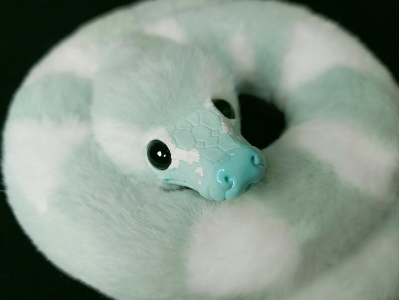 May include: A plush snake toy coiled in a circle, featuring soft mint green and white faux fur. The head is crafted from a smooth, light blue material with a textured scale pattern, large black glossy eyes, and a defined snout with nostrils.