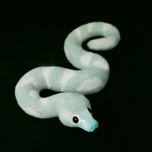 May include: A plush snake toy featuring a soft, fuzzy texture with alternating bands of light mint green and white. The snake has a smooth, solid mint green head with two dark, glossy eyes and a small, sculpted snout, curled into an S-shape.