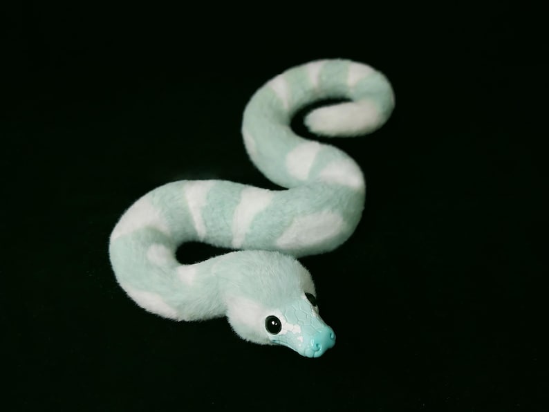 May include: A plush snake toy featuring a soft, fuzzy texture with alternating bands of light mint green and white. The snake has a smooth, solid mint green head with two dark, glossy eyes and a small, sculpted snout, curled into an S-shape.