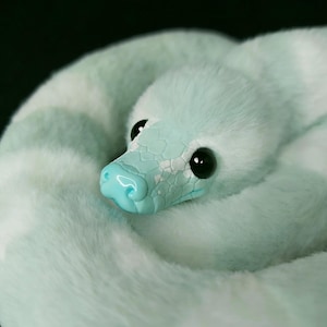 May include: A plush snake toy featuring a soft, light mint green faux fur body with white patches. The head is crafted from a smooth, glossy mint green material with a textured scale pattern and large, round, black plastic eyes.