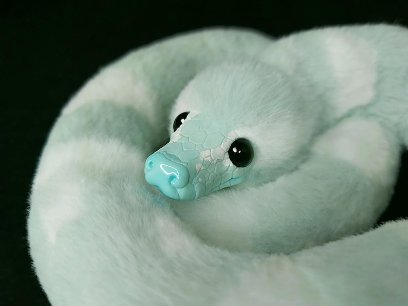 May include: A plush snake toy featuring a soft, light mint green faux fur body with white patches. The head is crafted from a smooth, glossy mint green material with a textured scale pattern and large, round, black plastic eyes.