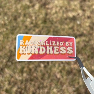 May include: A rectangular sticker with the text "RADICALIZED BY KINDNESS" in a retro font. The sticker has a white border and a colorful design with blue, orange, red, and white stripes.