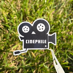 May include: A black and white sticker in the shape of a vintage film camera. The word "CINEPHILE" is printed in white block letters below the camera. The sticker is held by tweezers against a background of green grass.