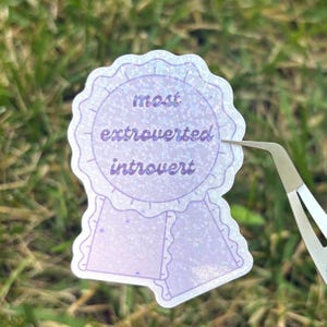 May include: A light purple sticker with the words "most extroverted introvert" in a circular design, resembling a ribbon. The sticker has a glittery finish and is being held by tweezers against a backdrop of green grass.