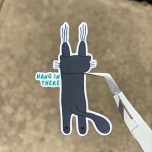 以下が含まれることがあります： ぶら下がっているポーズの漫画の猫が描かれたステッカー。猫は濃いグレーで、白いアクセントがあり、「HANG IN THERE」の文字が緑と白で書かれています。ステッカーはピンセットで持たれています。