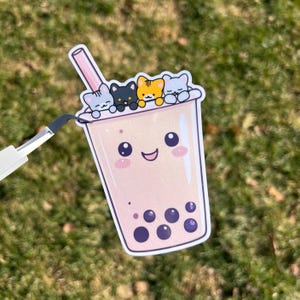 May include: A sticker featuring a cute cartoon boba tea cup with a smiling face and four cat characters on top. The boba tea is pink with dark purple boba pearls. A pink straw is visible. The sticker is on a green grassy background.