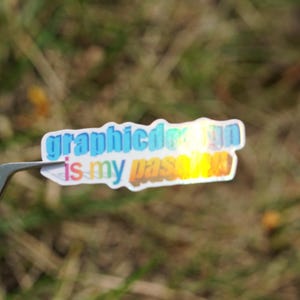 May include: A colorful sticker with the text "graphic design is my passion" in a gradient of blue, yellow, and orange. The sticker is held by a pair of silver tweezers against a blurred green background.