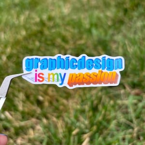 May include: A sticker with the text "graphicdesign is my passion" in blue, yellow, and orange. The sticker is held by tweezers against a blurred green grass background.