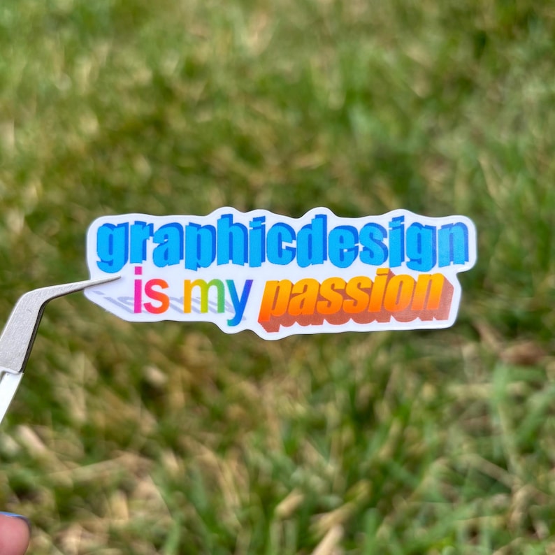 May include: A sticker with the text "graphicdesign is my passion" in blue, yellow, and orange. The sticker is held by tweezers against a blurred green grass background.
