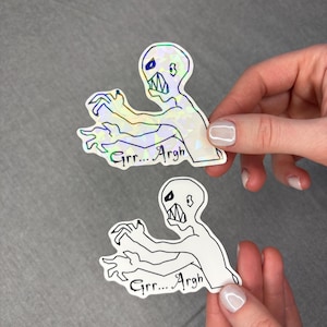 May include: Two stickers featuring a cartoon zombie with the text "Grrr... Argh". One sticker has a holographic finish, while the other is a black and white line drawing. The zombie has a wide mouth and outstretched arms.