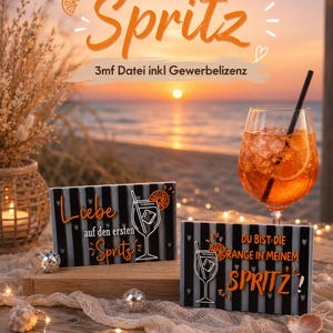 Op de afbeelding: Twee rechthoekige zwart-wit gestreepte bordjes met oranje en witte tekst en afbeeldingen van een cocktailglas met een schijfje sinaasappel. Het linkerbordje toont Liebe auf den ersten Spritz, het rechterbordje toont DU BIST DIE ORANGE IN MEINEM SPRITZ!
