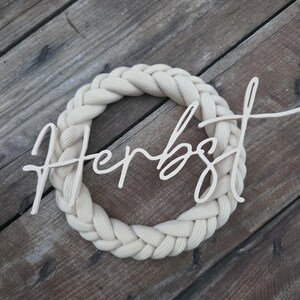 May include: A circular wreath made of thick, cream-coloured braided fabric. Centred across the wreath is a light-coloured wooden cutout featuring the word "Herbst" written in a cursive, flowing script font with a smooth, matte finish.