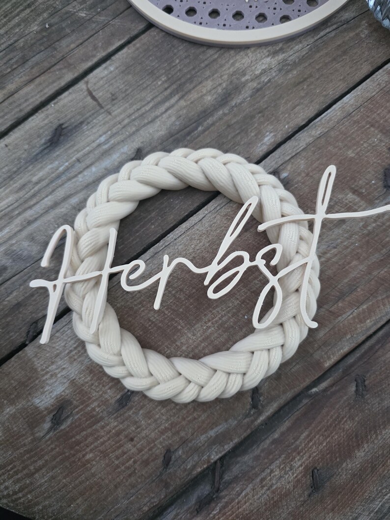 May include: A circular wreath made of thick, cream-coloured braided fabric. Centred across the wreath is a light-coloured wooden cutout featuring the word "Herbst" written in a cursive, flowing script font with a smooth, matte finish.
