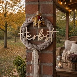 May include: A circular wreath made of braided beige fabric featuring a wooden cutout script that reads "Herbst" across the centre. The top is decorated with dried yellow and red flowers, and the bottom features hanging cream-coloured lace ribbons.
