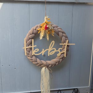 May include: A braided taupe fabric wreath featuring a wooden cutout script word "Herbst" across the centre. The top is decorated with a small cluster of dried yellow and red flowers, while the bottom features a hanging cream-coloured lace ribbon.
