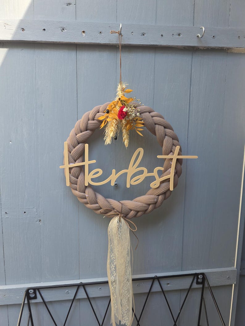 May include: A braided taupe fabric wreath featuring a wooden cutout script word "Herbst" across the centre. The top is decorated with a small cluster of dried yellow and red flowers, while the bottom features a hanging cream-coloured lace ribbon.