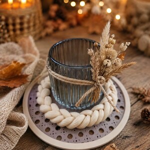 May include: A ribbed dark blue glass candle holder wrapped in twine with a small bouquet of dried beige grasses and flowers. The holder sits inside a thick, braided cream-coloured yarn ring on a circular beige base with a perforated pattern.