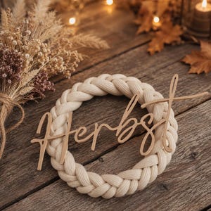 May include: A circular wreath made of thick, braided cream-coloured fabric cord. A light brown wooden cutout script word "Herbst" is attached across the centre of the braided ring, featuring a cursive font style with a long, extended crossbar.