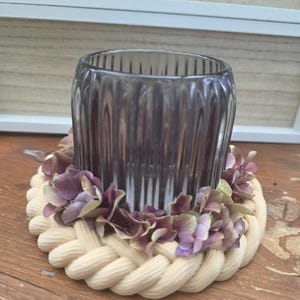May include: A cylindrical, ribbed, dark purple glass candle holder sits inside a circular base. The base is constructed from a thick, cream-coloured braided rope and is decorated with small, dried purple hydrangea flower petals.