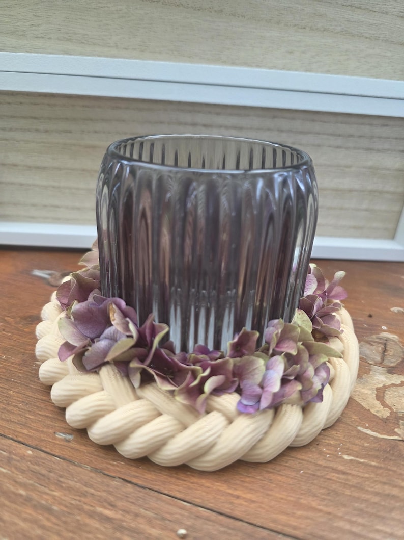 May include: A cylindrical, ribbed, dark purple glass candle holder sits inside a circular base. The base is constructed from a thick, cream-coloured braided rope and is decorated with small, dried purple hydrangea flower petals.
