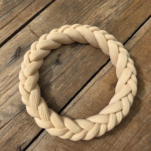 May include: A circular ring featuring a braided rope design with a light beige, matte finish. The object is constructed with a continuous, interlocking pattern that mimics the appearance of woven strands, creating a textured, uniform surface.