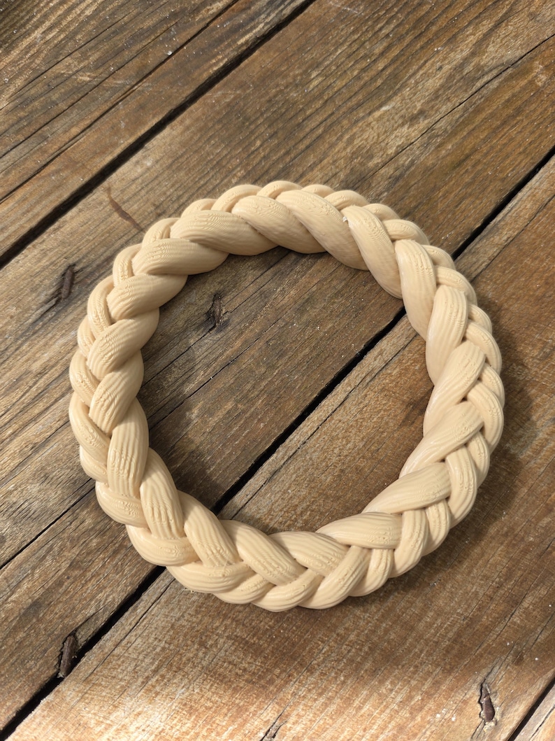 May include: A circular ring featuring a braided rope design with a light beige, matte finish. The object is constructed with a continuous, interlocking pattern that mimics the appearance of woven strands, creating a textured, uniform surface.