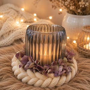 May include: A cylindrical, ribbed, dark grey glass candle holder sits inside a circular base made of thick, braided cream-coloured rope. A ring of dried, muted purple hydrangea petals is tucked between the glass holder and the rope base.