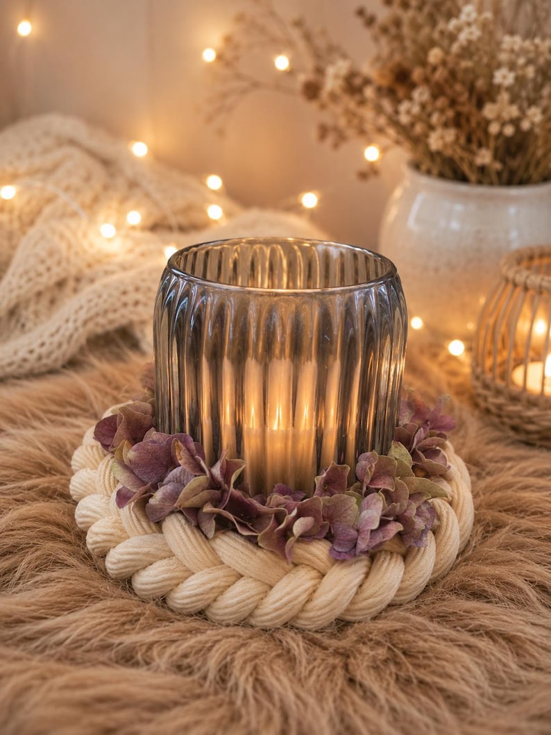 May include: A cylindrical, ribbed, dark grey glass candle holder sits inside a circular base made of thick, braided cream-coloured rope. A ring of dried, muted purple hydrangea petals is tucked between the glass holder and the rope base.