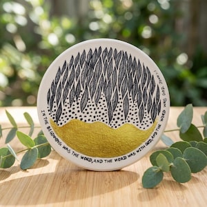 Könnte beinhalten: Ein runder, cremefarbener Keramikuntersetzer mit einem schwarz-goldenen Design. Die Oberseite zeigt ein Muster aus schwarzen Blättern und Sternen über einem goldfarbenen, strukturierten Band. Der Text "IN THE BEGINNING WAS THE WORD, AND THE WORD WAS WITH GOD, AND THE WORD WAS GOD. JOHN 1:1" ist aufgedruckt.
