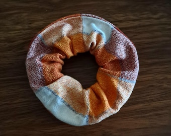 Op de afbeelding: Een cirkelvormige stoffen haarelastiek met een ruitpatroon in tinten oranje, wit en bruin. Het accessoire heeft een geplooide, gerimpelde textuur die wordt gecreëerd door een interne elastische band, met een geweven textielconstructie.