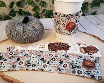 Op de afbeelding: Twee stoffen koffiebekerhoezen met een bloemenpatroon en een bruin uilontwerp. Een hoes heeft een bruine knoop met een uilontwerp. De hoezen liggen op een houten oppervlak met een grijze gebreide pompoen.