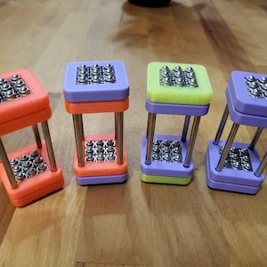May include: Four colorful, geometric desk toys with a square top and bottom connected by silver metal rods. The tops and bottoms are orange, purple, and green, each holding small, silver spheres. The toys are arranged on a wooden surface.
