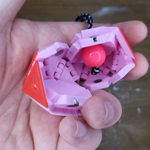 May include: A pink and red geometric container with a pink sphere inside. The container is open, revealing the sphere and interior details. A black and white cord is attached to the top. The object is held in a hand.