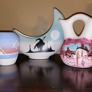 May include: Three ceramic vases with Southwestern designs. The vase on the left is blue, pink, and white with geometric patterns. The centre vase is white and blue with a bird silhouette. The vase on the right is white with a desert landscape.
