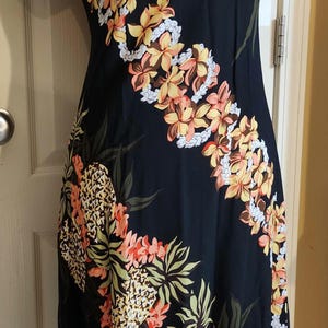 May include: A black short-sleeved dress featuring a repeating diagonal pattern of orange and yellow tropical flowers, white lei-style floral garlands, and pineapple motifs. The garment has a round neckline and a straight, knee-length hemline.