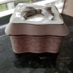 May include: White porcelain trinket box with a raised white bow on the lid. The box has a scalloped edge and a delicate floral pattern.