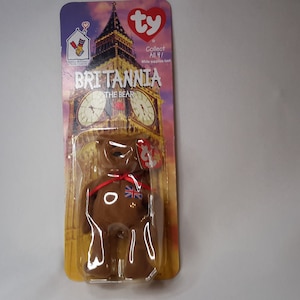 May include: A brown Beanie Baby plush toy named Britannia the Bear, wearing a red ribbon and a Union Jack patch. The toy is packaged in a plastic bag with a cardboard backing featuring a picture of Big Ben and the text "Collect All 4!" and "Britannia the Bear".