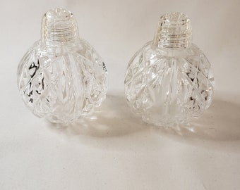May include: Two clear, glass salt and pepper shakers. Each shaker has a round, diamond-cut body and a ridged, screw-on top. The shakers are displayed on a white surface. These decorative shakers are ideal for adding a touch of elegance to any dining table.