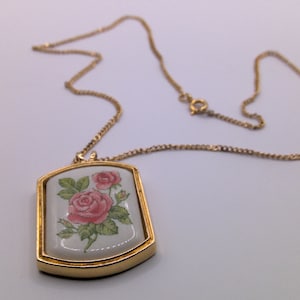 May include: A gold chain necklace with a rectangular pendant featuring a white porcelain cameo with a pink rose design.