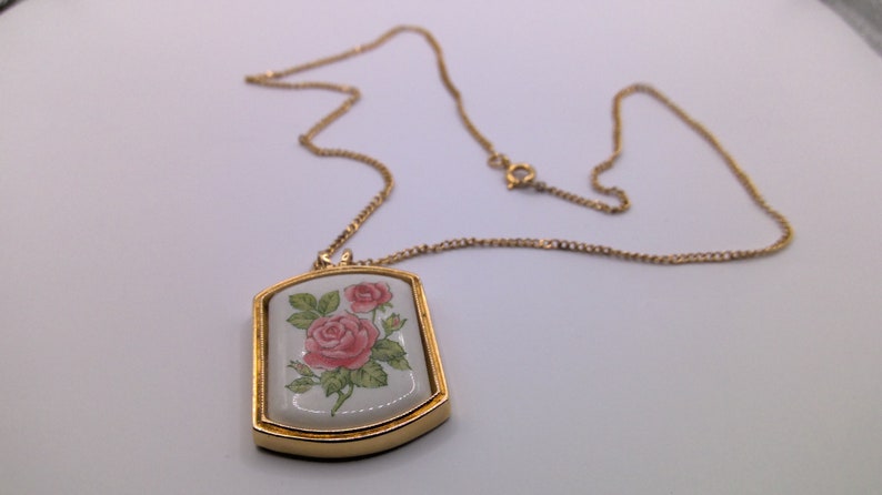 May include: A gold chain necklace with a rectangular pendant featuring a white porcelain cameo with a pink rose design.