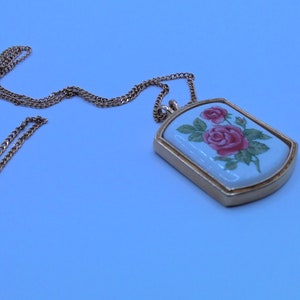 May include: A gold chain necklace with a rectangular pendant featuring a white porcelain design of pink roses.