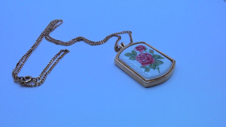 May include: A gold chain necklace with a rectangular pendant featuring a white porcelain design of pink roses.