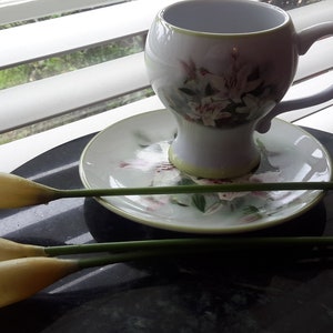 May include: A white teacup and saucer set with a floral design, resting on a dark surface. Two yellow calla lilies with green stems are placed near the cup. The cup has a handle and a matching floral pattern.