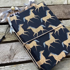 May include: A rectangular journal covered in black fabric featuring a repeating pattern of tan horses with white manes and tails. The journal is secured with a thin tan elastic band and includes a silver ribbon, a gold button, and a black tassel.