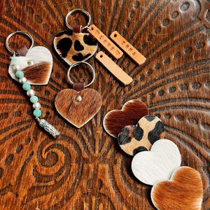 May include: Heart-shaped keychains and charms in various textures and patterns, including cowhide and leopard print. Some keychains have beaded accents. Small rectangular charms are engraved with the words "love" and "emma".