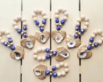 May include: Set of eight gold heart-shaped napkin rings with oyster shells inside. Each ring is made of wooden beads with blue and gold accents.