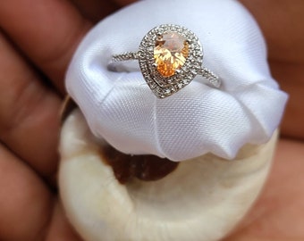 May include: A pear-shaped ring with a large, orange-brown gemstone surrounded by a halo of smaller, clear gemstones. The ring is set in silver and has a simple band.